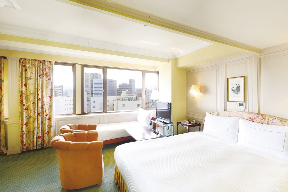 Une chambre d’hôtel avec un grand lit, un bureau avec une télévision, un canapé et une vue sur le paysage urbain grâce à de grandes fenêtres.