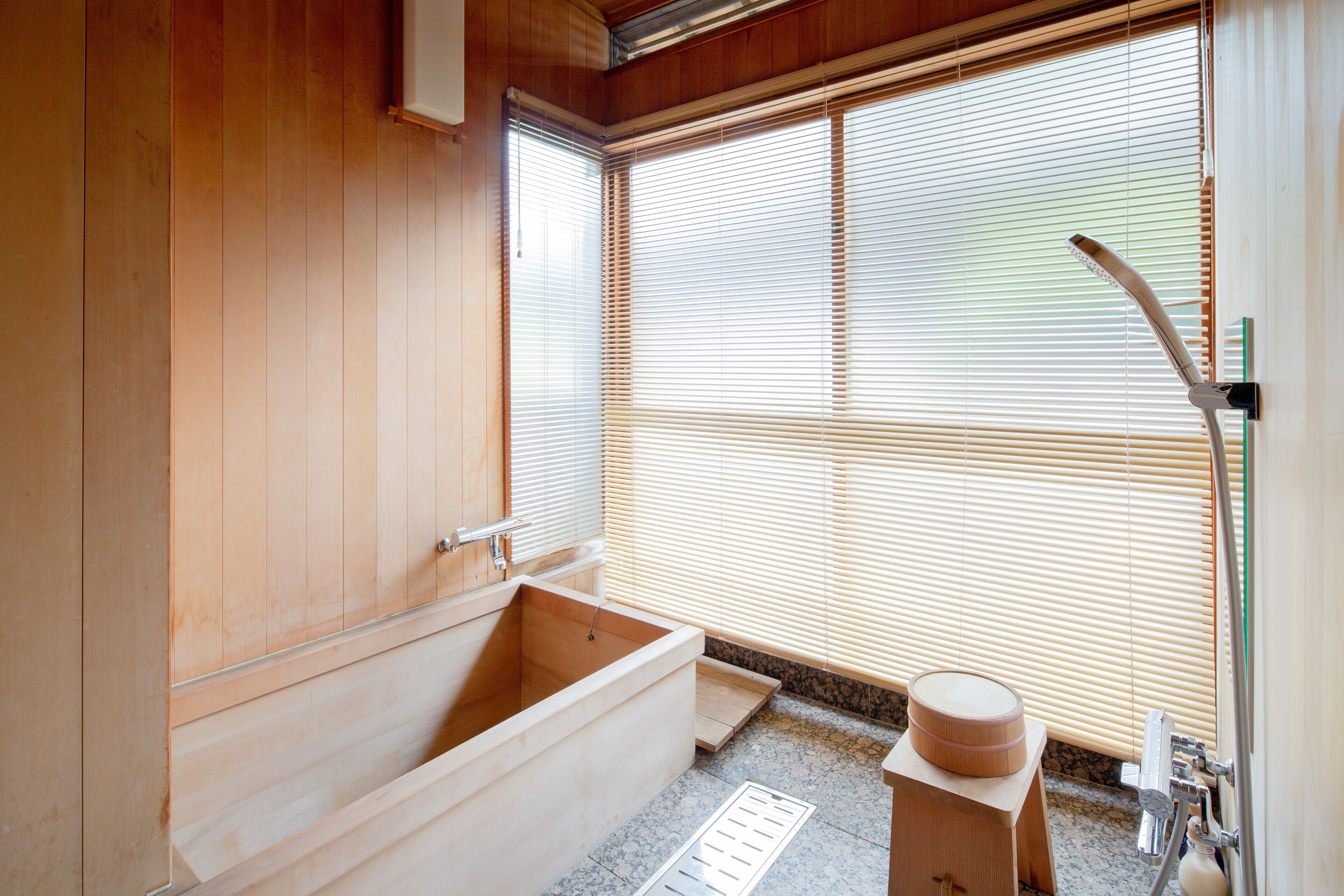 Un baño de estilo japonés tradicional con una bañera de madera, un taburete pequeño de madera y una ventana con persianas.
