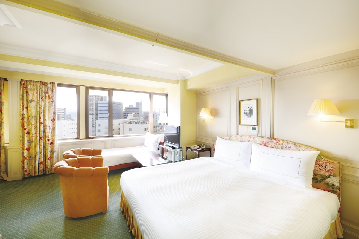 Habitación de hotel con una cama grande, dos camas más pequeñas, un sofá, un escritorio, un televisor y una ventana con vista a la ciudad.