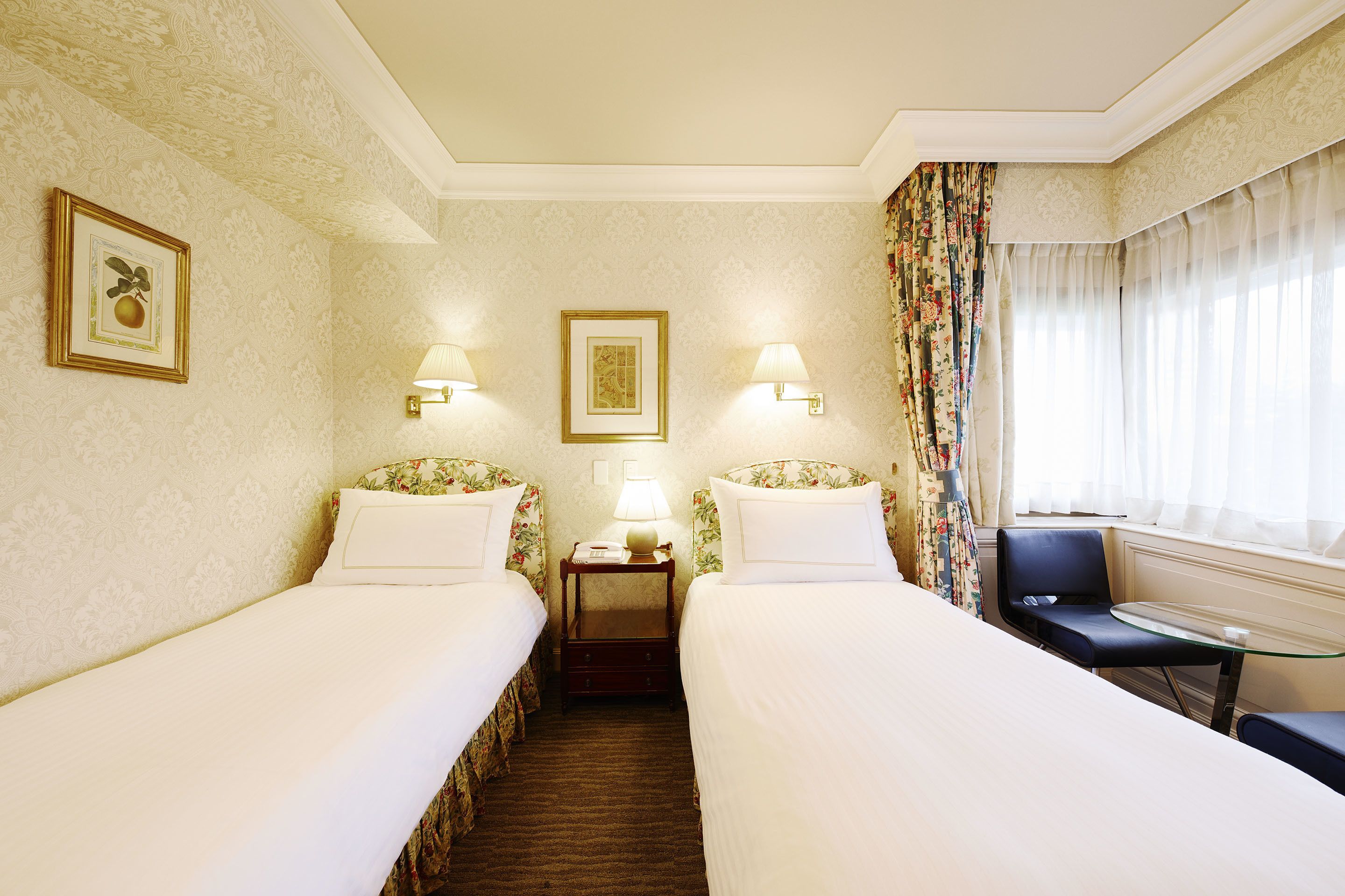 A hotel room with two single beds, a small table, two nightstands with lamps, and a chair.