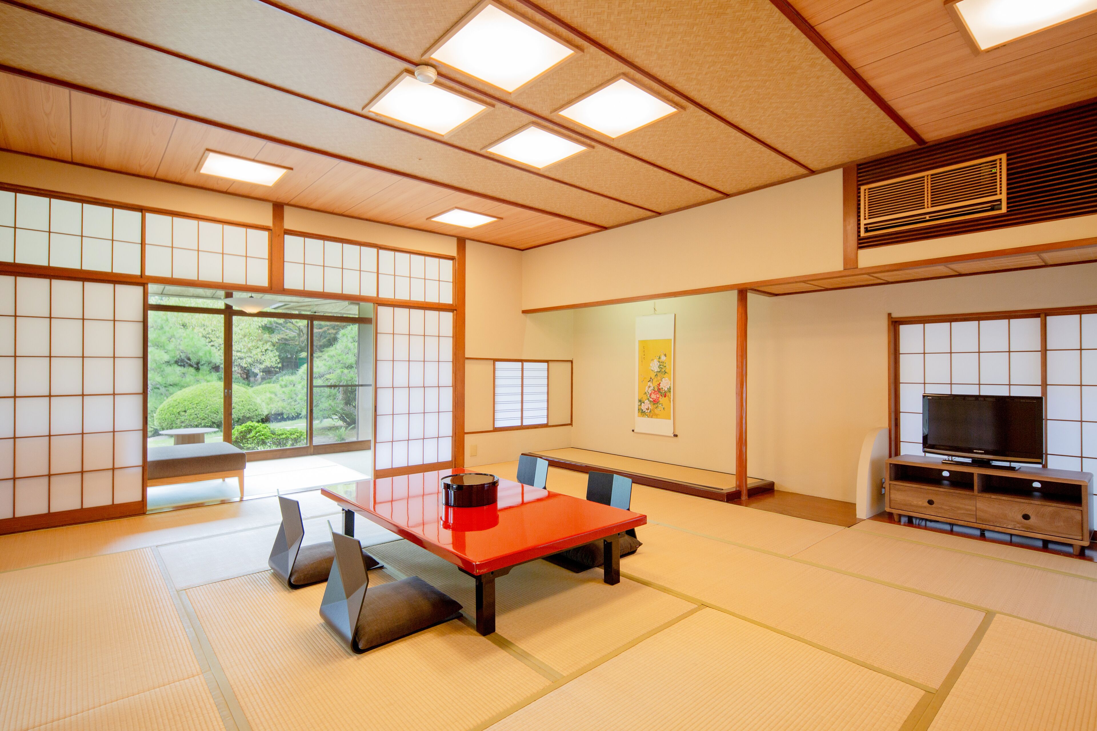 A traditional Japanese room with tatami flooring, a low table with cushions, a television, and sliding doors leading to a garden view.