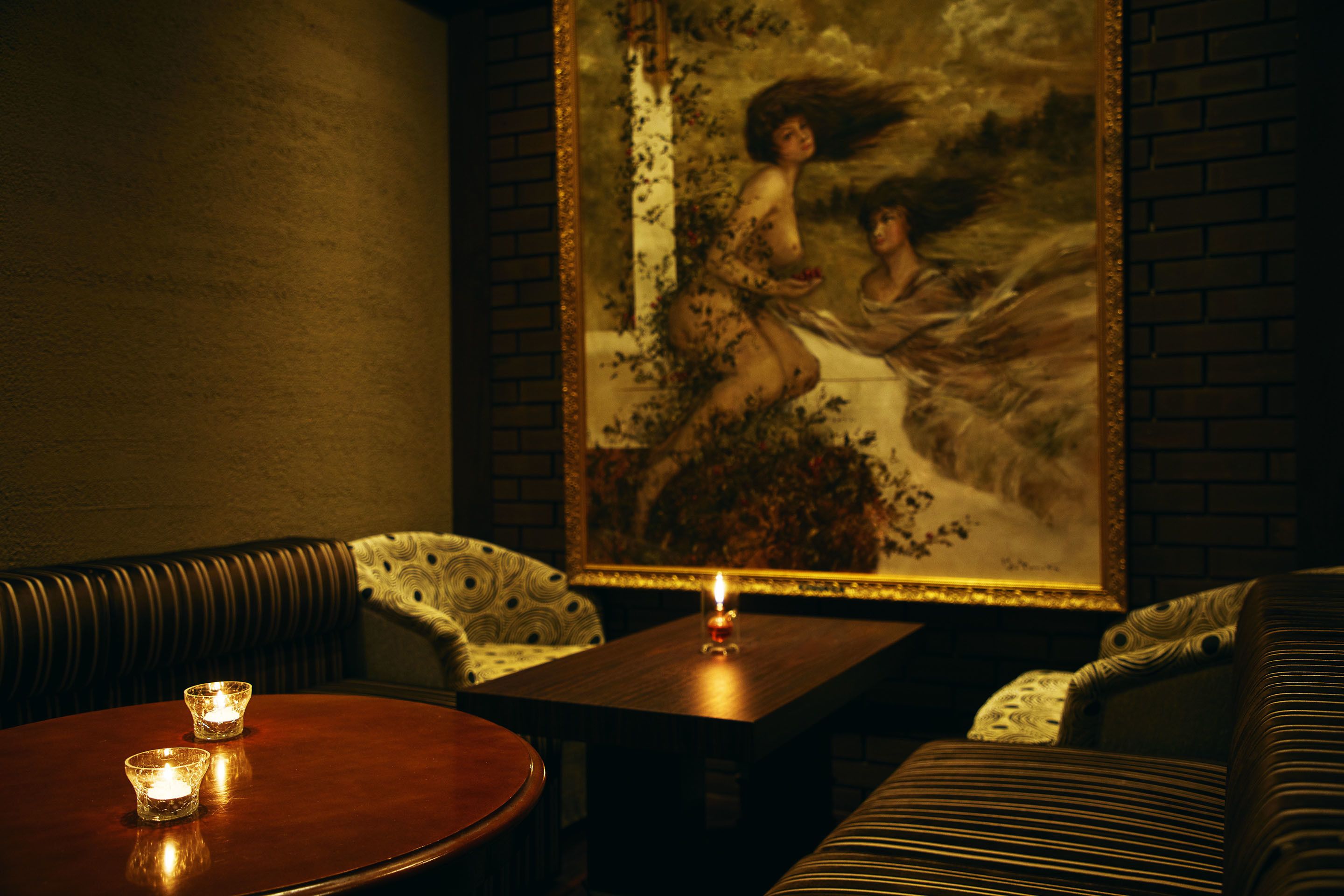 A dimly lit room with a round wooden table, two candles, and a large framed painting on the wall.