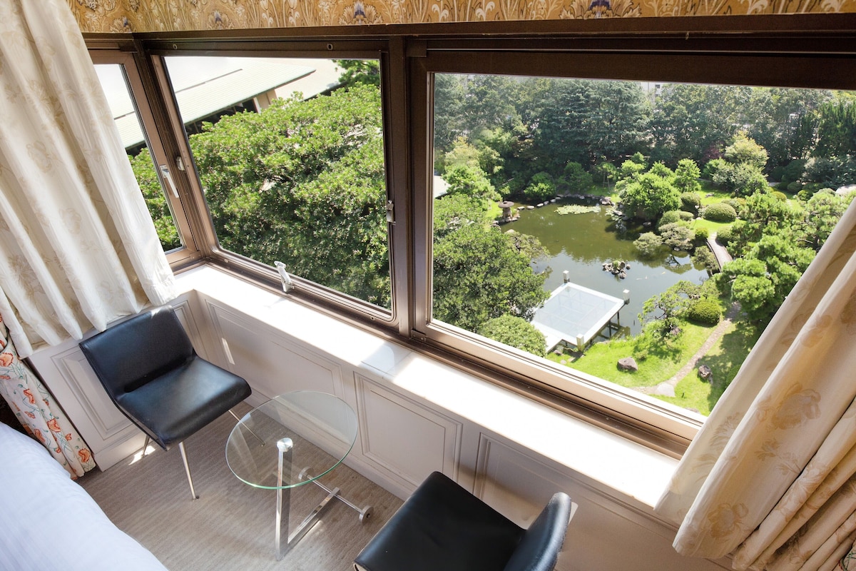 Habitación con un amplio ventanal que ofrece vistas a un jardín exuberante y un estanque. Hay dos sillas negras y una pequeña mesa de cristal.