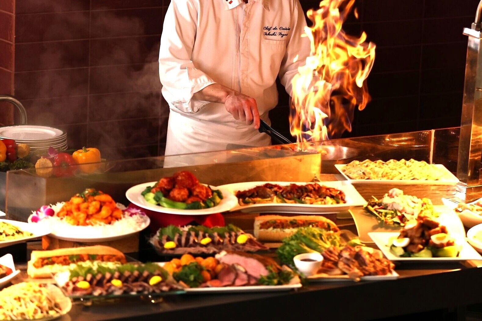 A chef cooking with flames, surrounded by a variety of dishes on a buffet table.