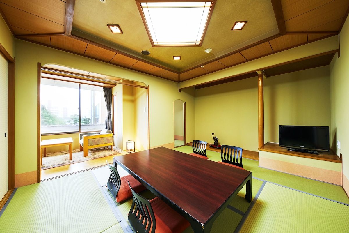 Una habitación con esteras de tatami, una mesa de madera, sillas, un televisor y un ventanal grande.