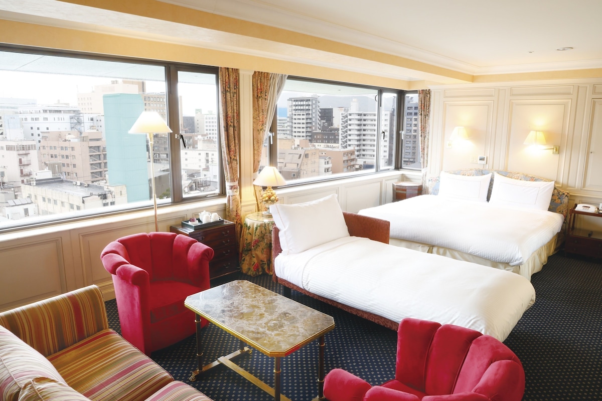 Un cuarto de hotel con una cama grande, dos sillones rojos, un sofá a rayas, una mesita y vistas a la ciudad a través de amplias ventanas.
