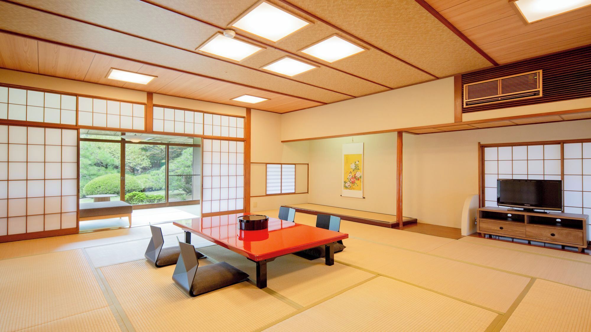 Ein traditioneller japanischer Raum mit Tatami-Bodenbelag, einem Tisch aus Tatami-Matten, einem Fernseher auf einem Holzständer und einer Rutschtür mit Blick auf Grünflächen.