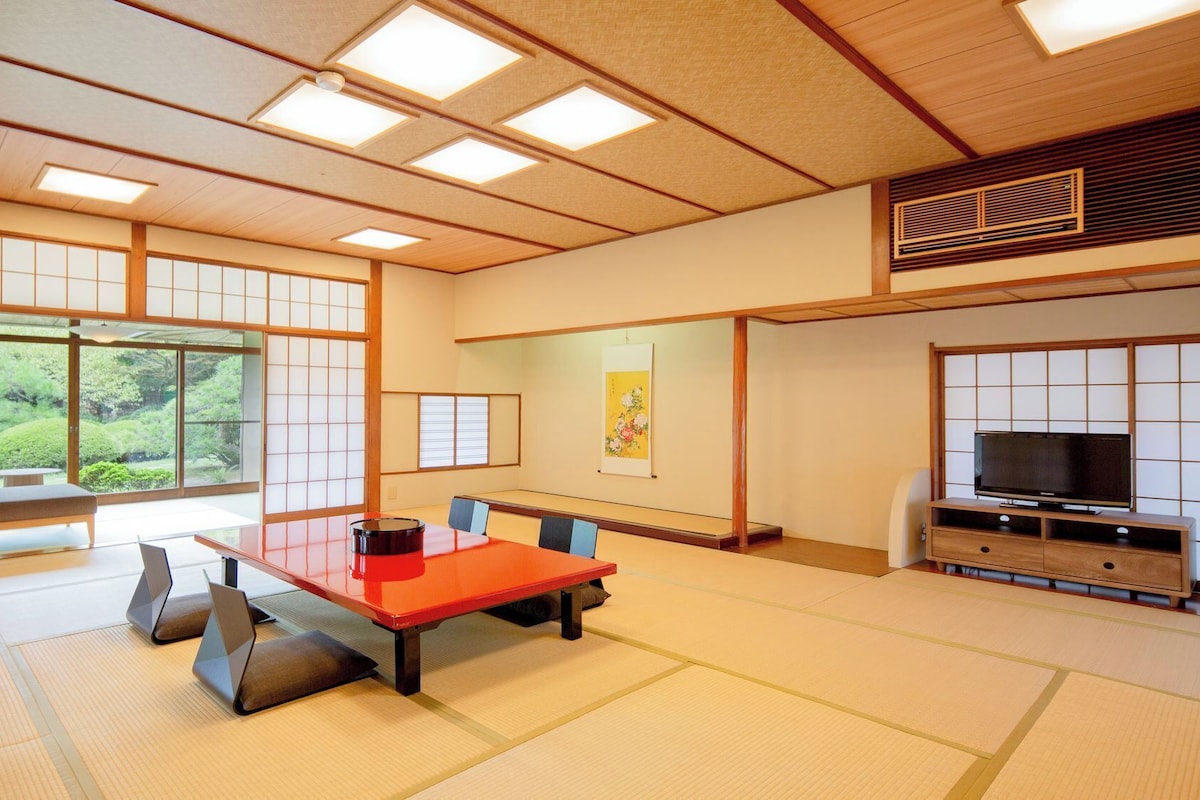 Una habitación tradicional japonesa con suelo de tatami, una mesa baja de tatami, un televisor sobre un soporte de madera y puertas correderas que dan a vistas de vegetación al exterior.