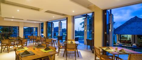 A spacious dining area with wooden tables and chairs, large windows offering views of the outdoors, and a pool area visible through the glass doors.