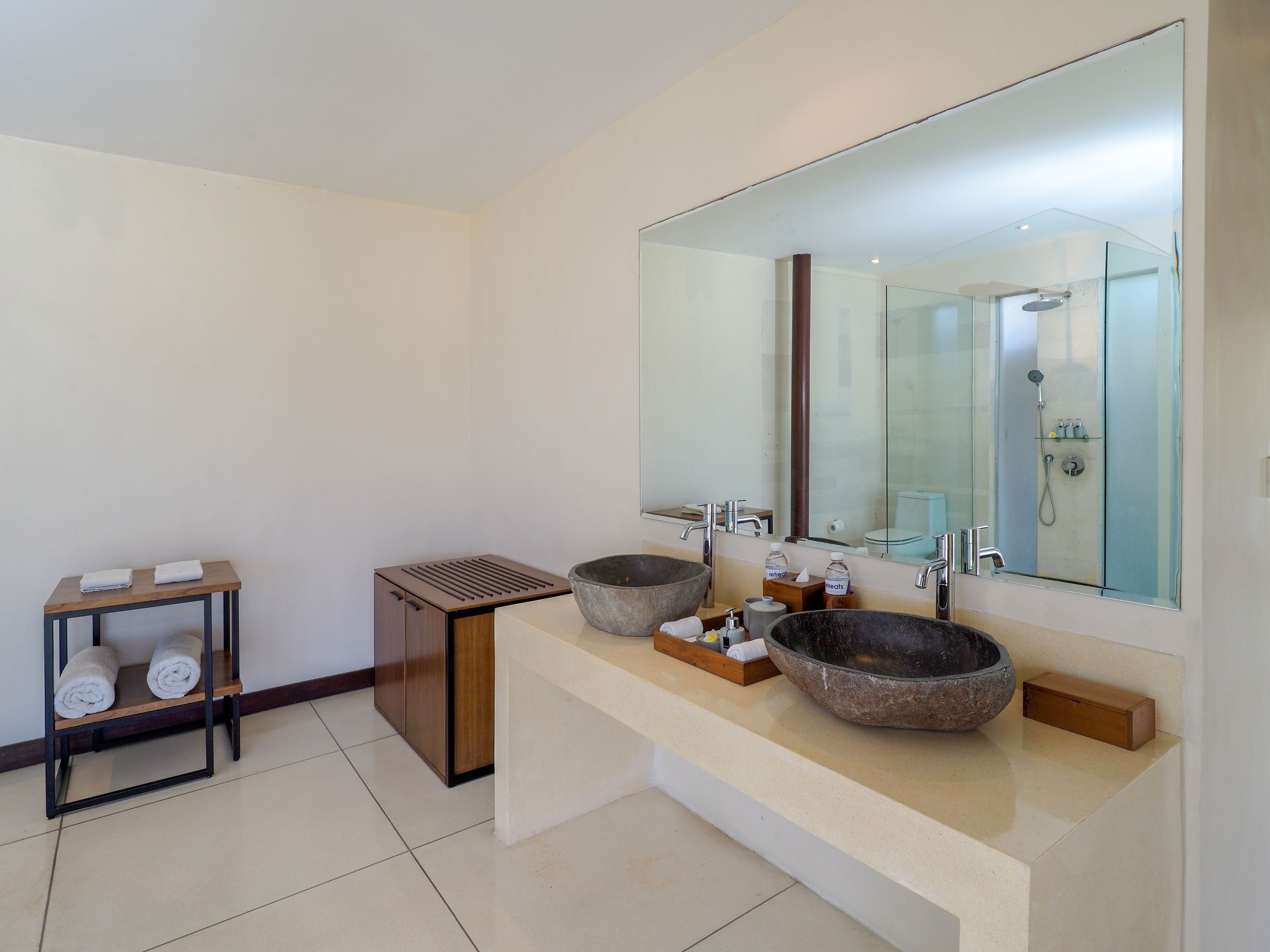 Un baño moderno con un espejo grande, dos lavamanos con cuencas amplias, un estante pequeño con toallas y una zona de ducha visible en el espejo.