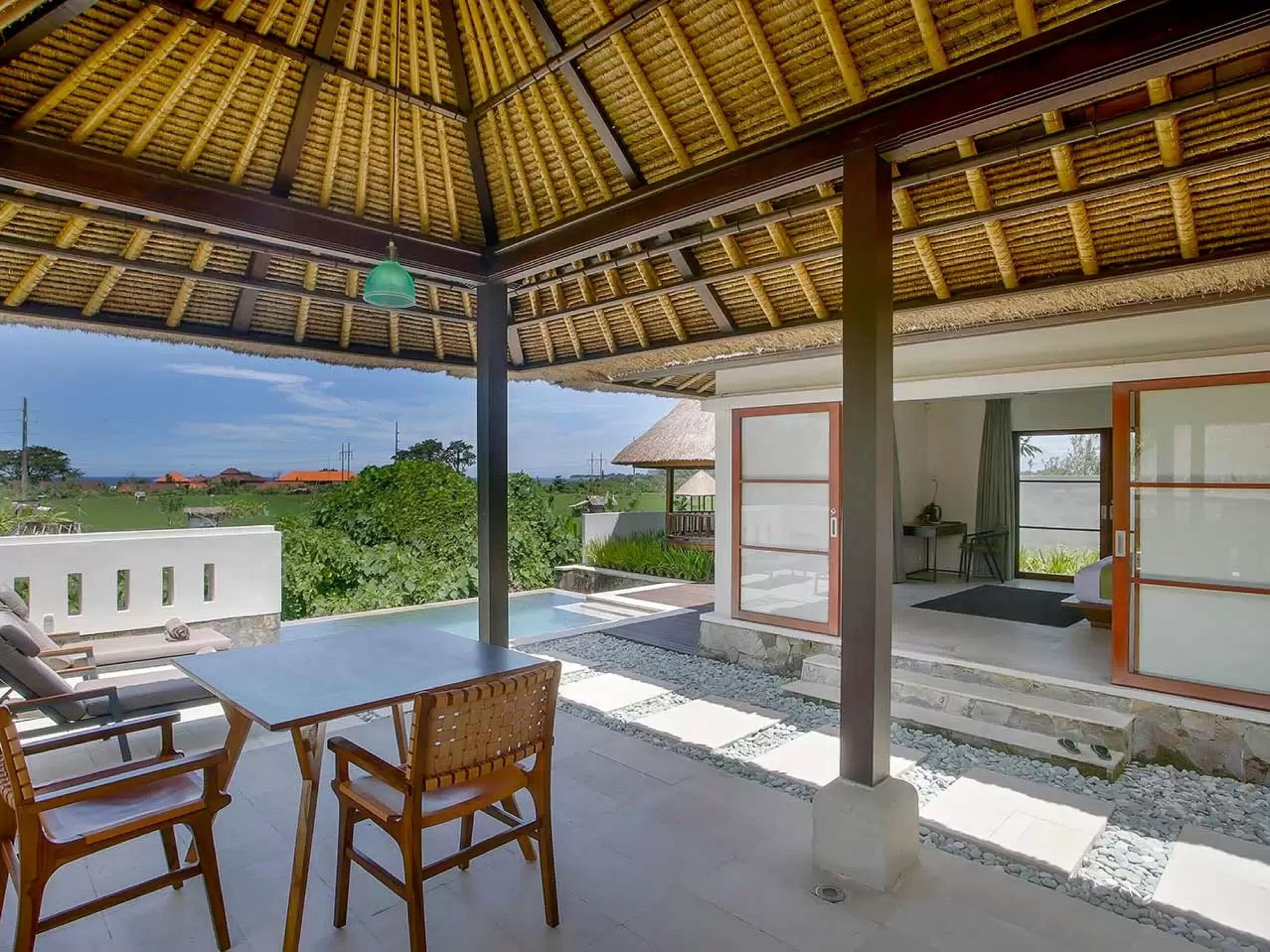 A covered outdoor seating area with a table and chairs, a glass door leading to a room, and a view of the surrounding landscape.