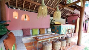 A cozy outdoor seating area with a wooden bench, wicker stools, and hanging lights. - Kuldesak Villas (Bandung)