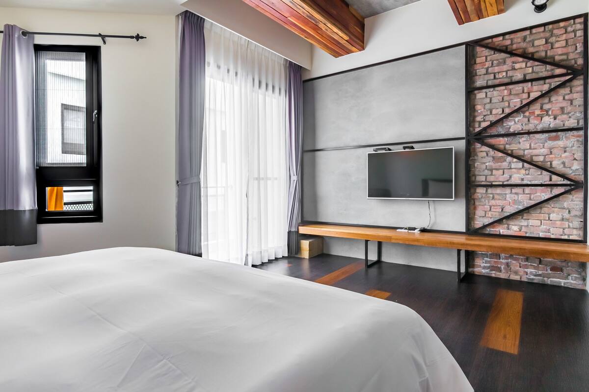Une chambre d’hôtel moderne avec un grand lit, un téléviseur fixé au mur en briques et un banc.