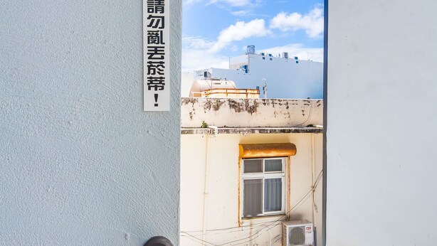 A narrow corridor with a sign on the wall, an air conditioning unit, and a window with a view of the sky and another building. - Warehouse Inn (Hengchun)