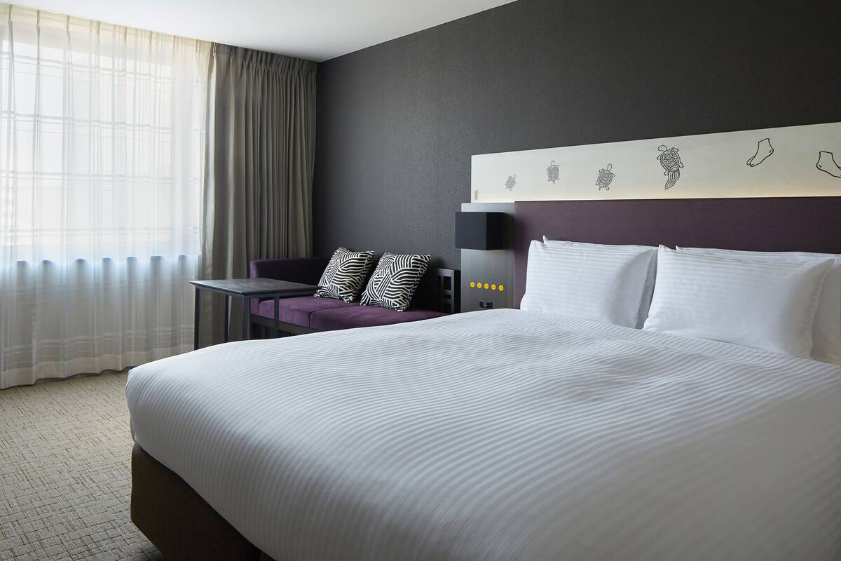 Habitación de hotel con una cama grande, una mesita pequeña, una silla morada y una pared con detalles decorativos.