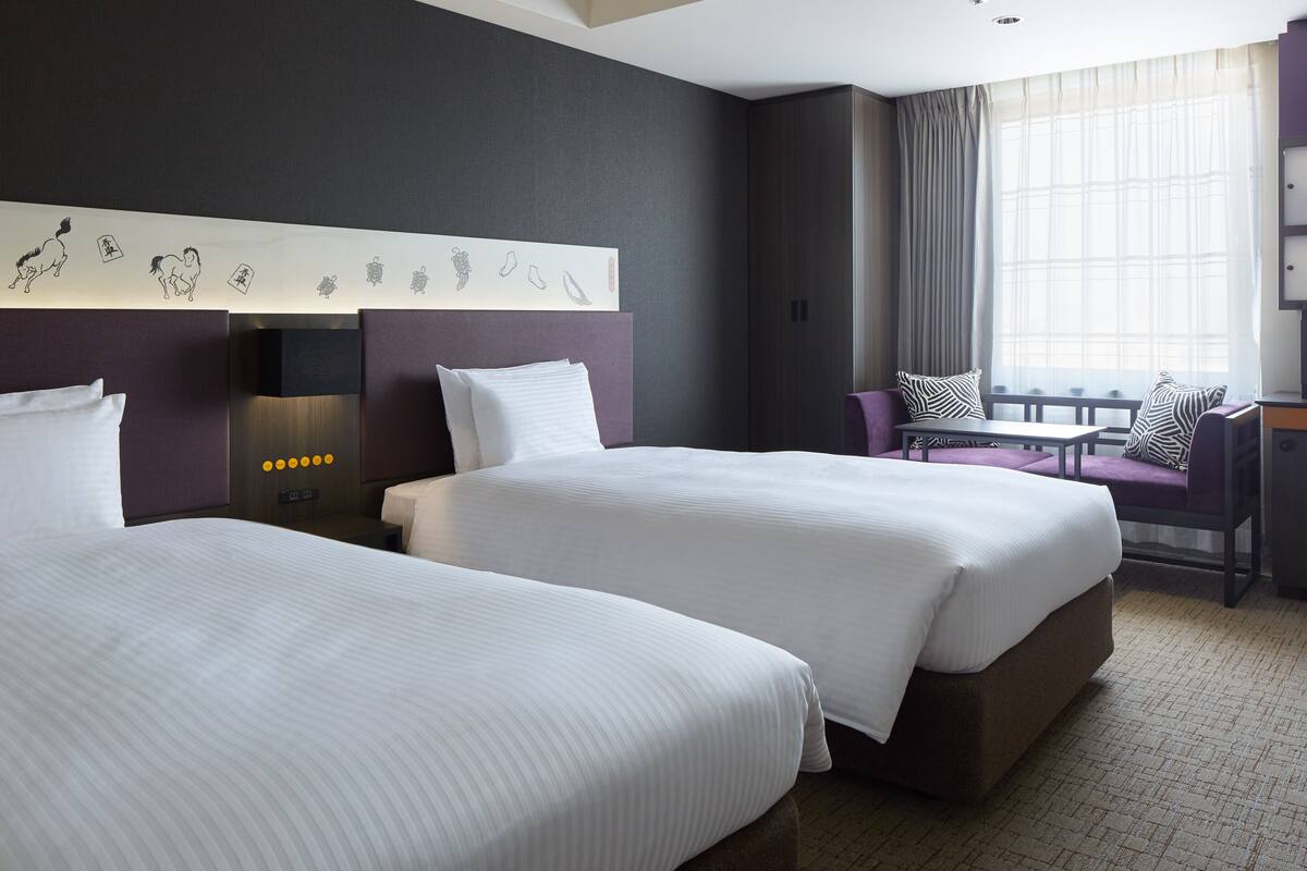 Habitación de hotel con dos camas, zona de estar con una silla y mesa, ventana grande con cortinas y una pared decorada con obras de arte.
