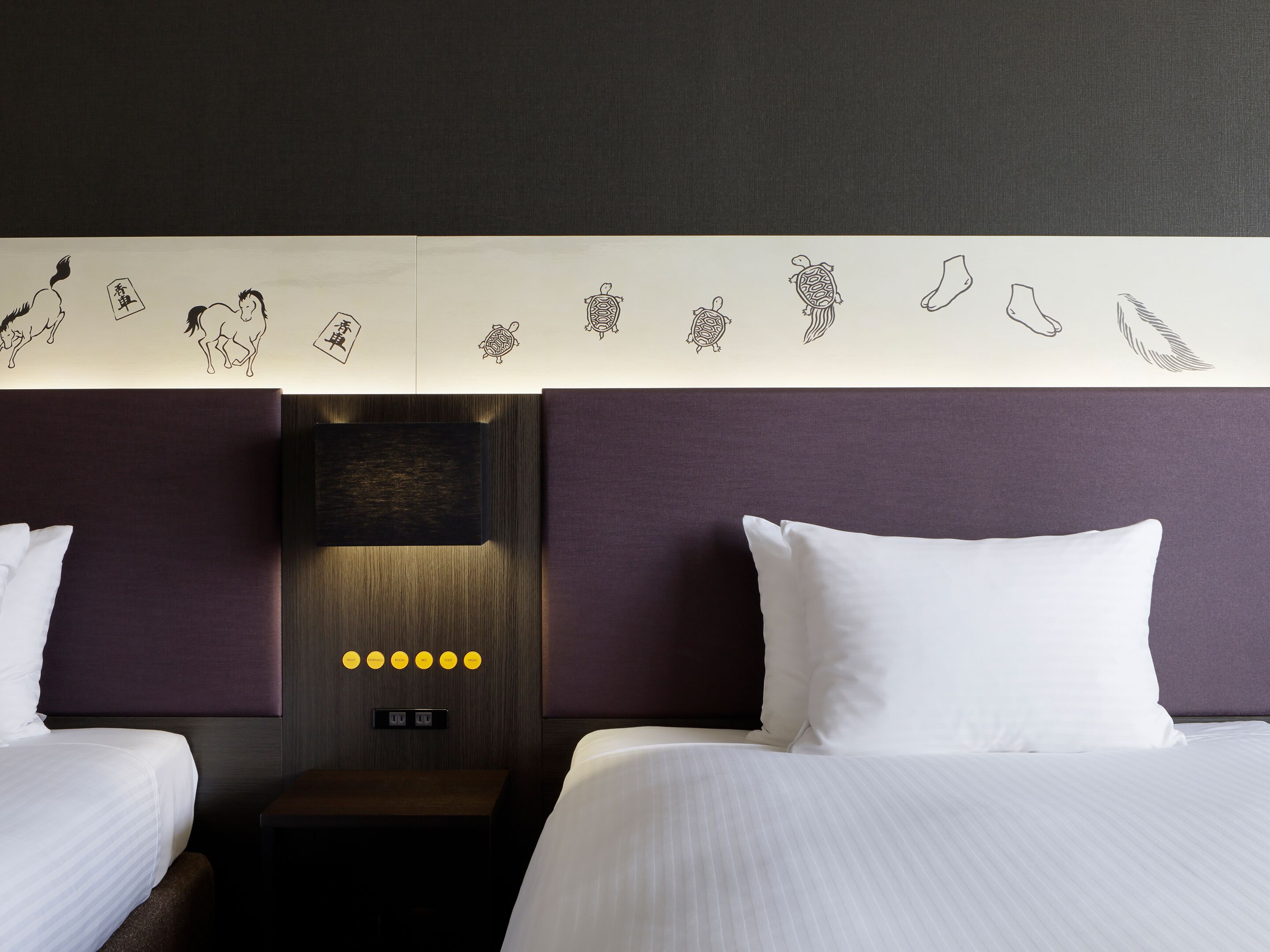 A hotel room with two beds, a headboard with wall art, a nightstand, and a control panel with buttons.