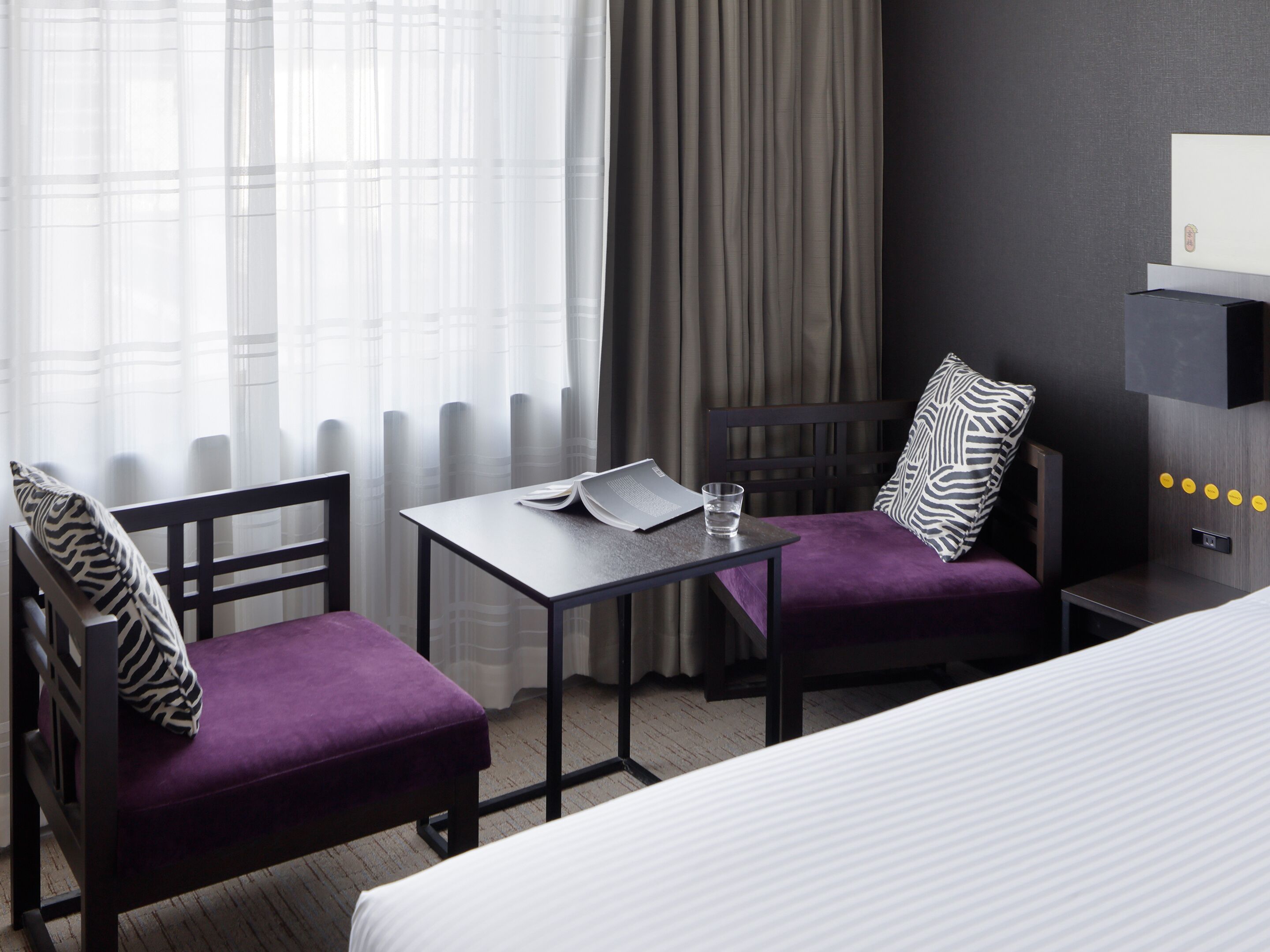 A hotel room with a bed, a desk with a chair, a purple ottoman, a zebra-patterned pillow, and a window with sheer curtains.