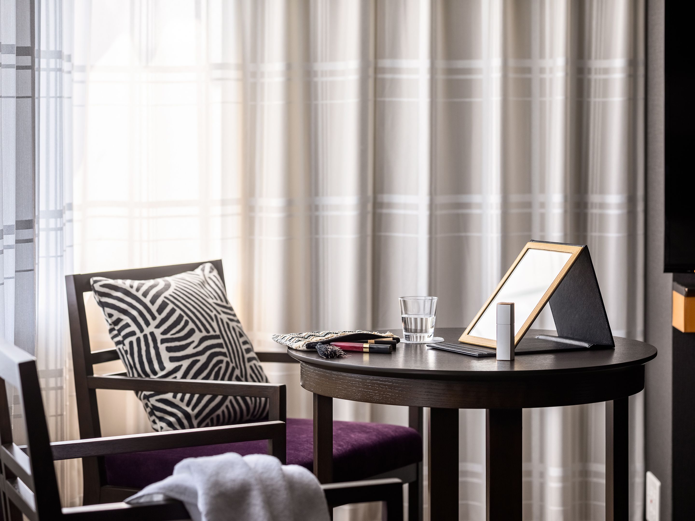 A hotel room with a round table, a chair with a zebra-patterned pillow, a glass of water, a framed picture, and a remote control.