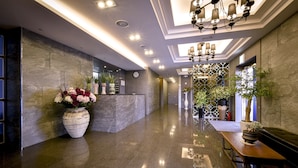 Un moderno vestíbulo de hotel con un mostrador de recepción de mármol, un gran jarrón de flores, un banco de madera y un aplique decorativo en el techo.