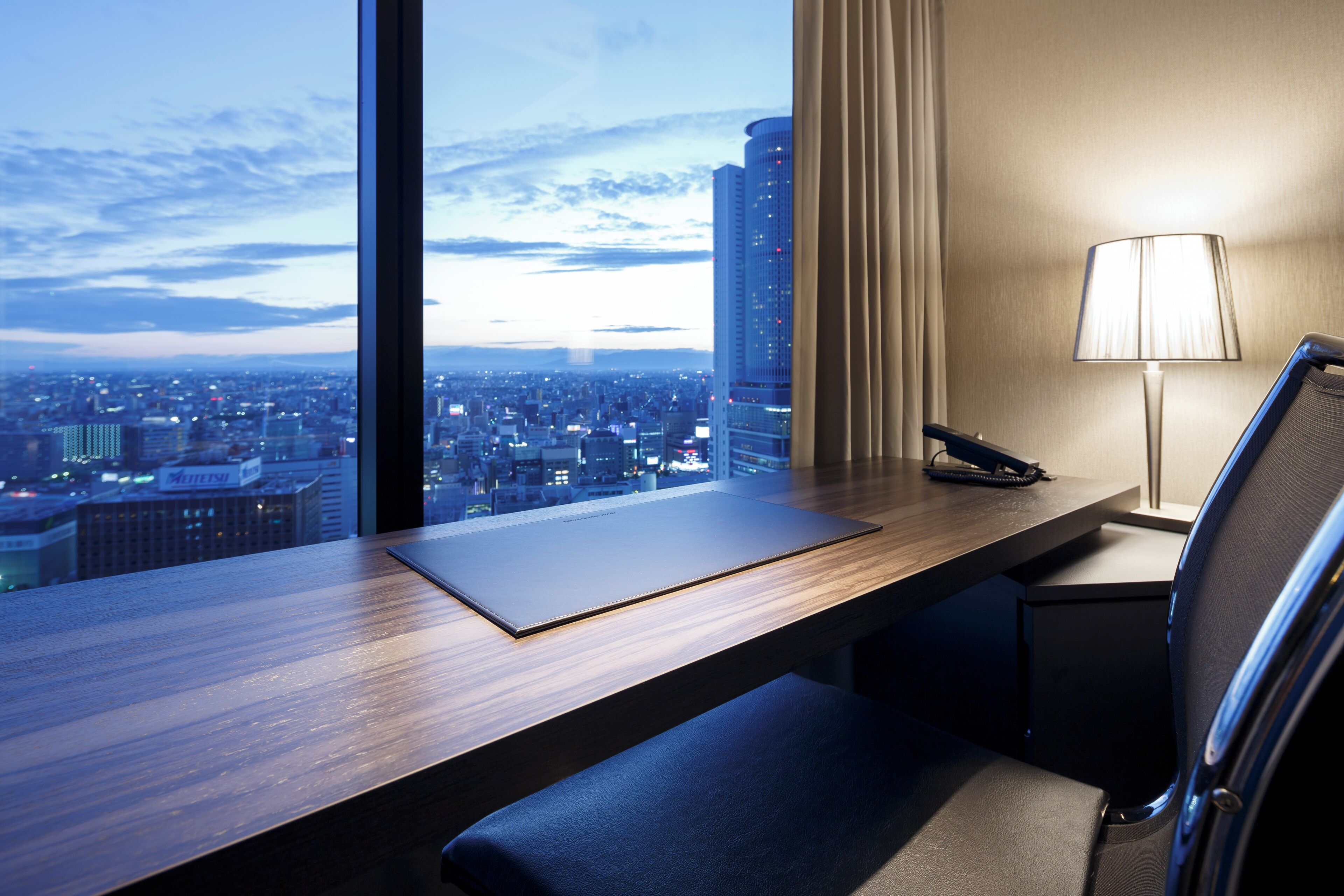 Quarto de hotel com escrivaninha, cadeira, abajur e vista para a cidade.