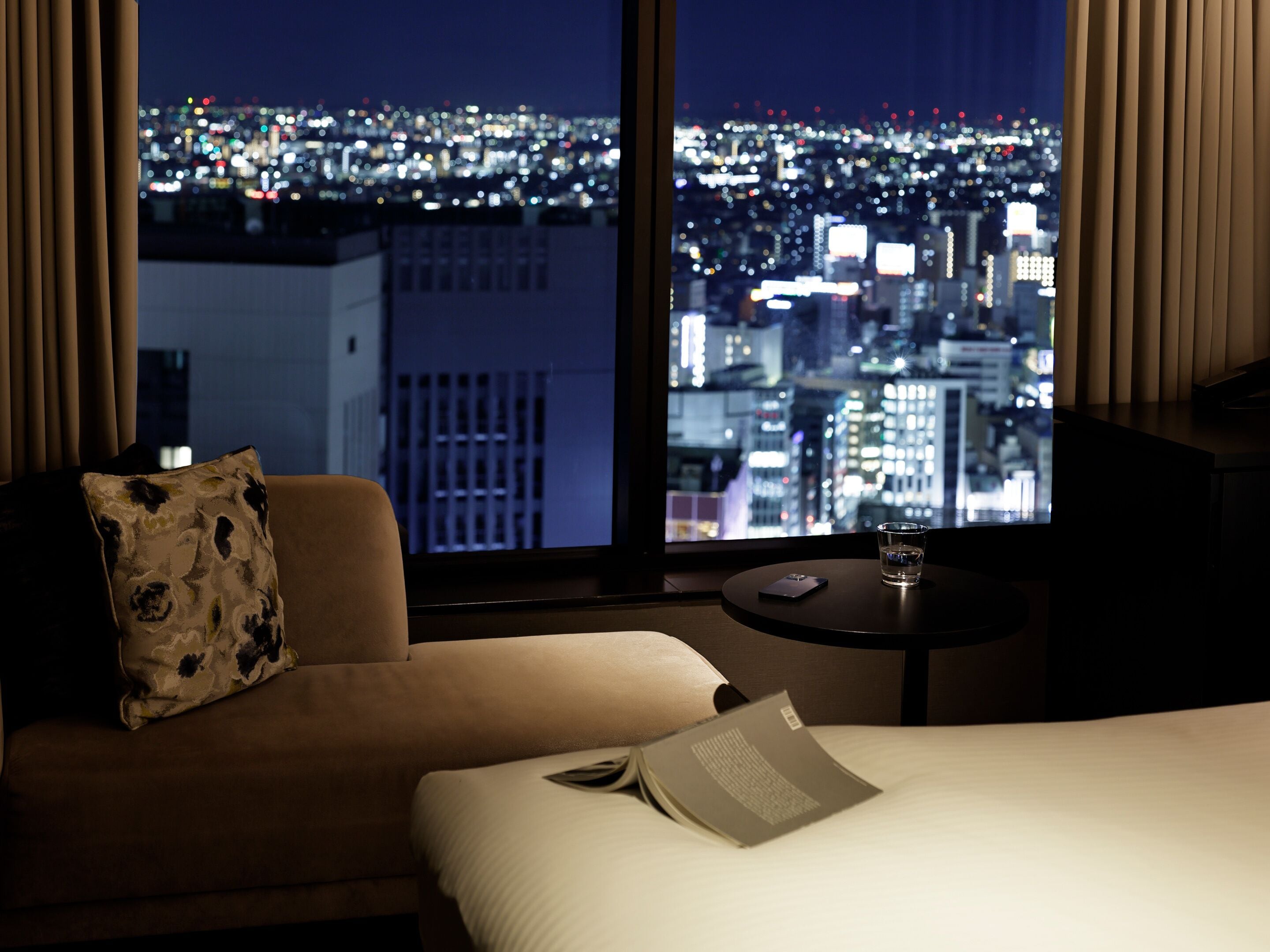 A hotel room with a bed, a sofa, a small table, and a view of a cityscape at night.