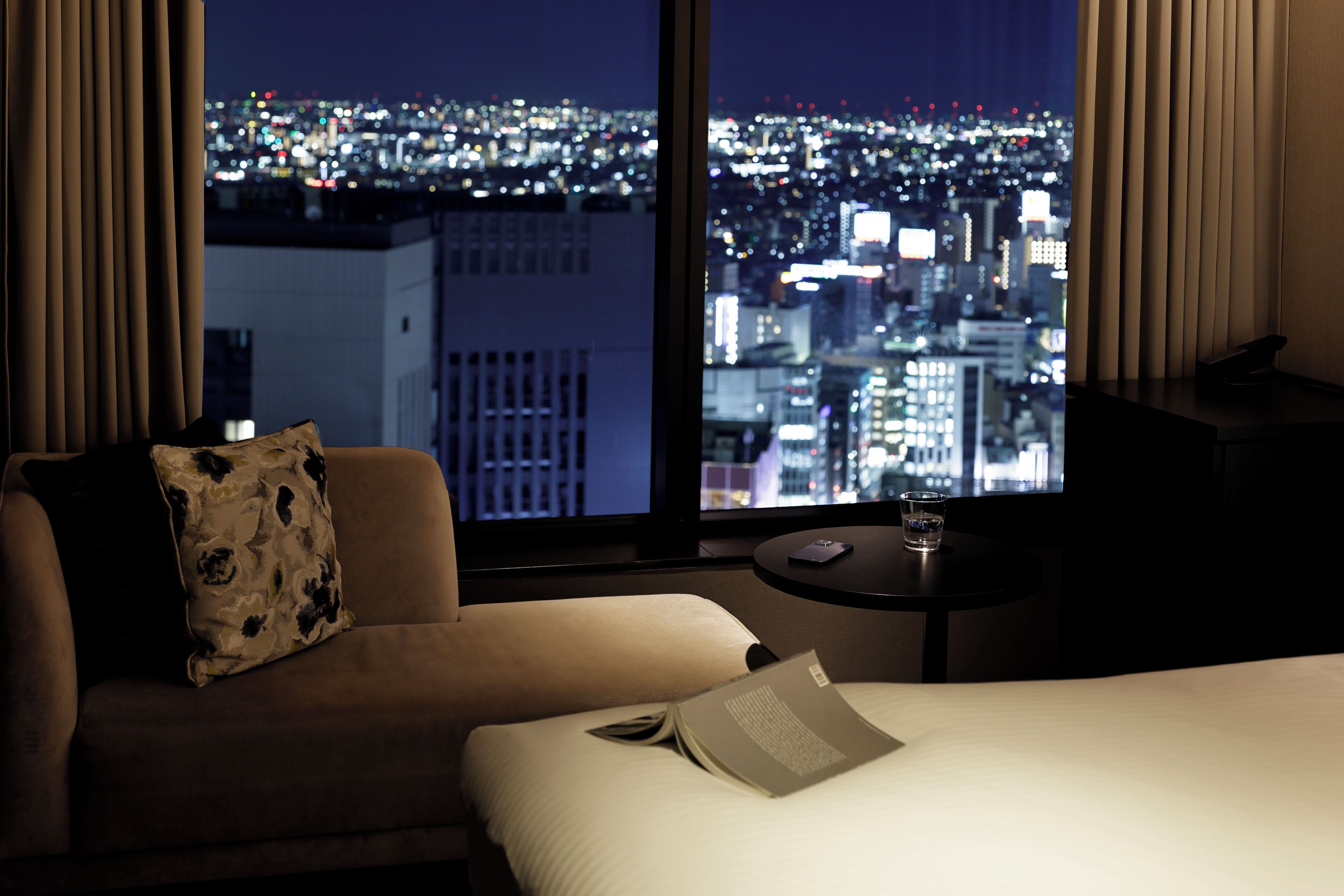 Habitación de hotel con cama, sofá, una mesita y vista a un paisaje urbano nocturno.