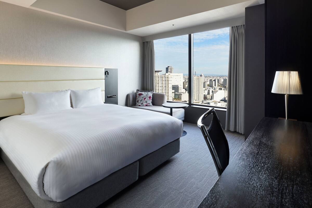 Une chambre d’hôtel avec un grand lit, un bureau, une chaise, une petite table et une vue sur la ville grâce à de grandes fenêtres.