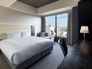 Ein Hotelzimmer mit einem großen Bett, einem Schreibtisch, einem Sessel, einem kleinen Tisch und Blick auf die Stadt durch große Fenster.