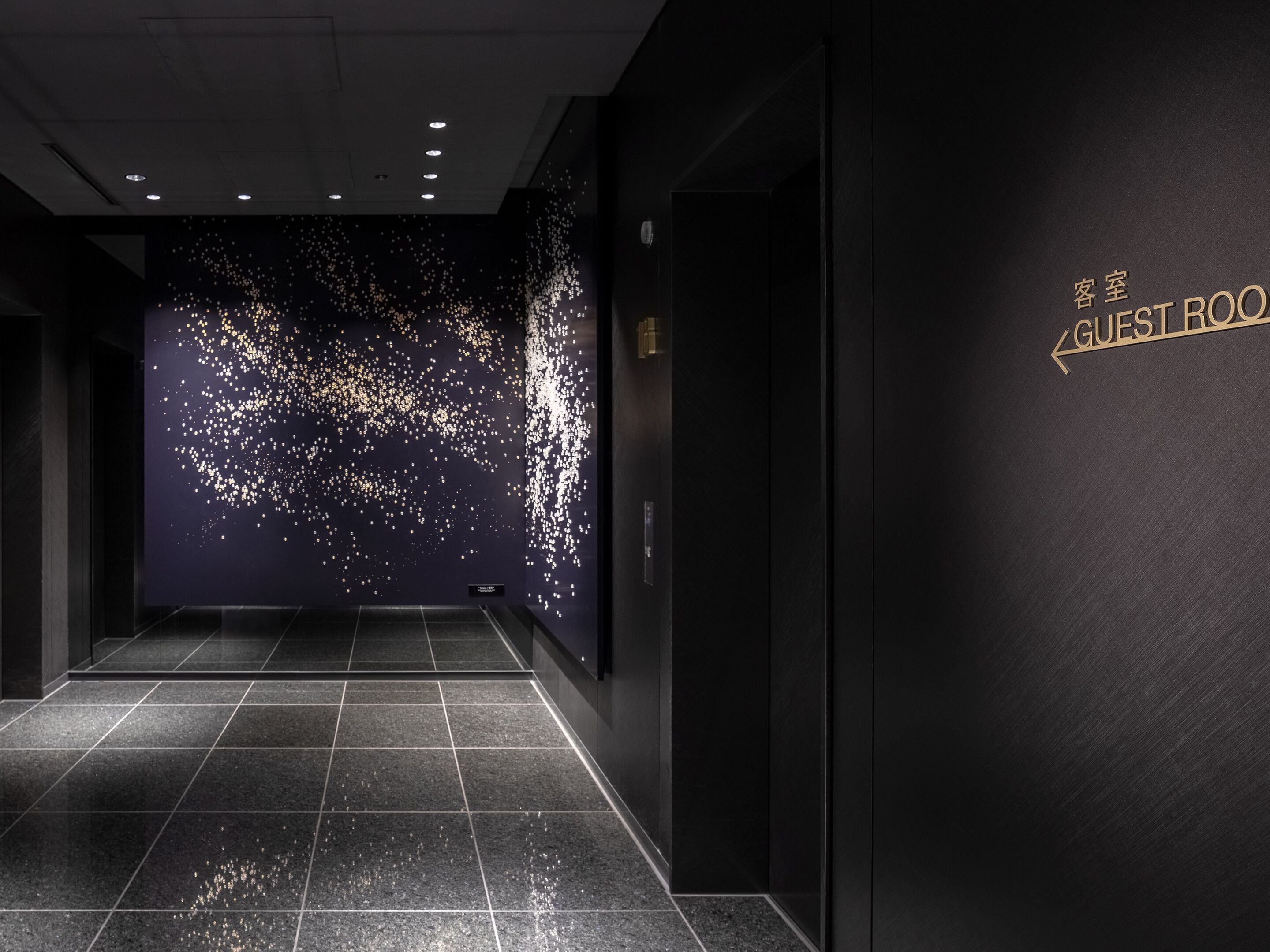 A modern hotel lobby with a dark floor, a wall with a 'Guest Room' sign, and a ceiling with recessed lighting.