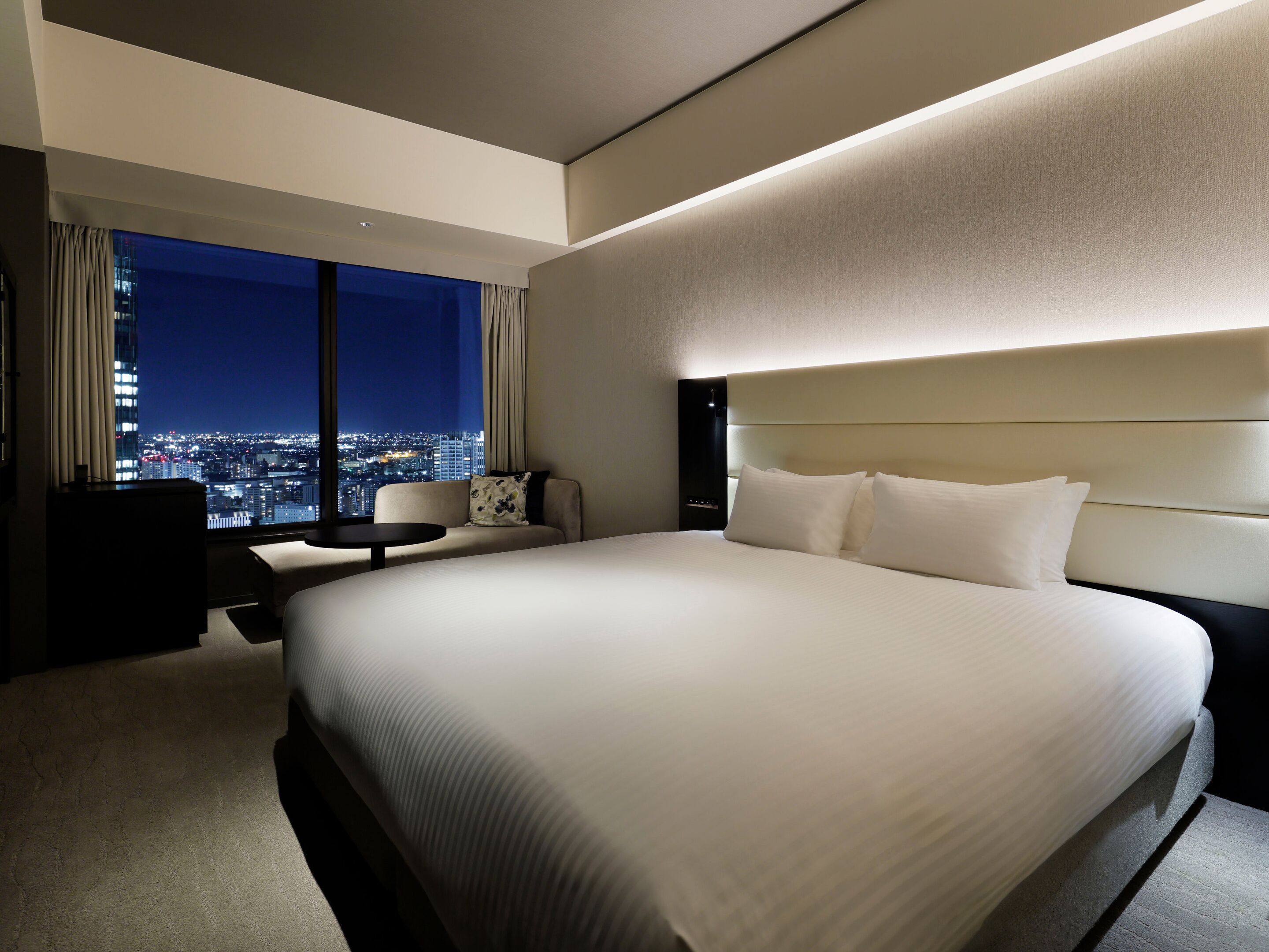 A modern hotel room with a large bed, a desk, a chair, and a view of a cityscape at night.