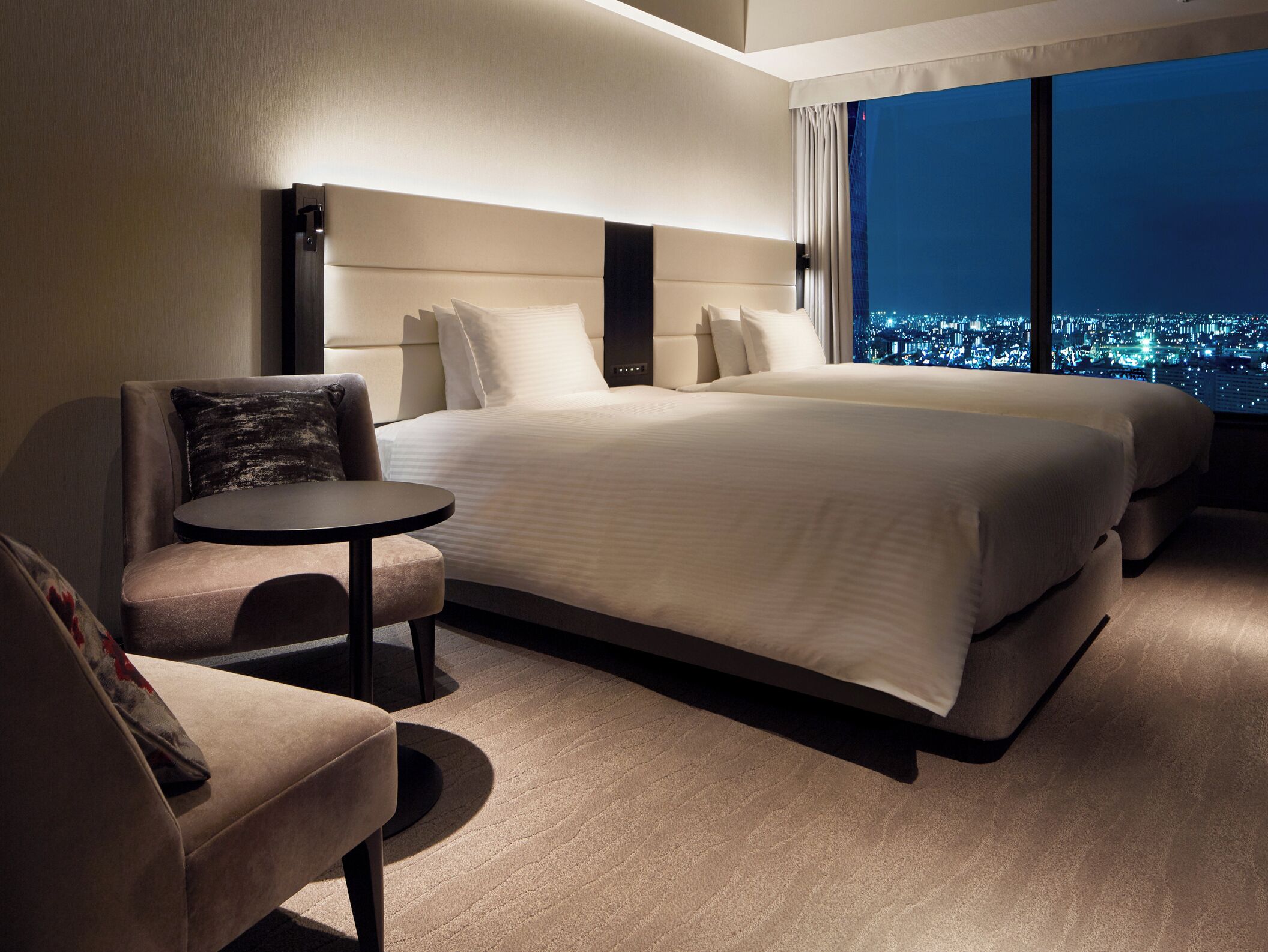 Habitación de hotel con una cama grande, una mesita, una silla y vistas a la ciudad de noche.