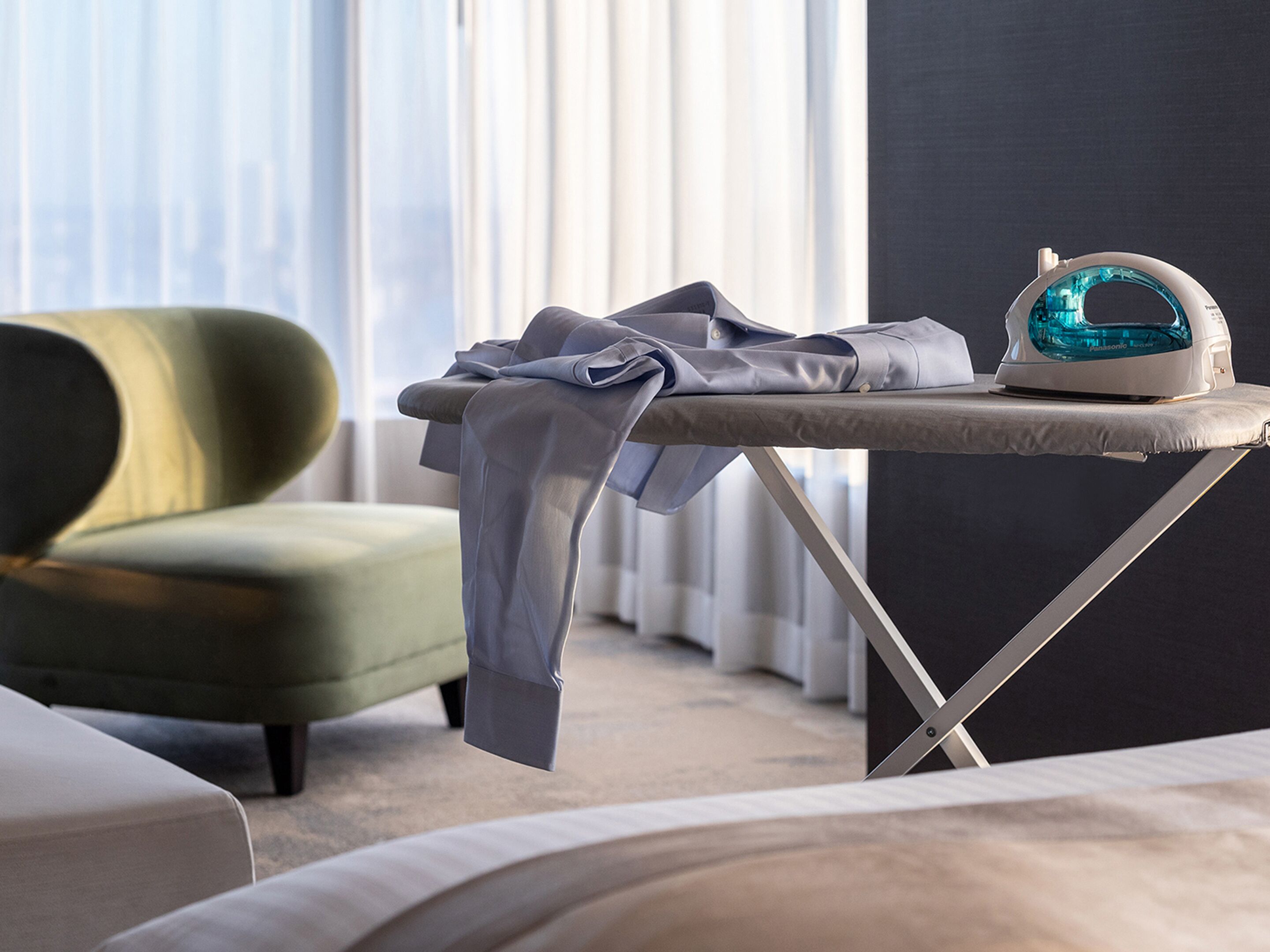A neatly made bed, a green armchair, and an ironing board with an iron in a modern room with large windows.