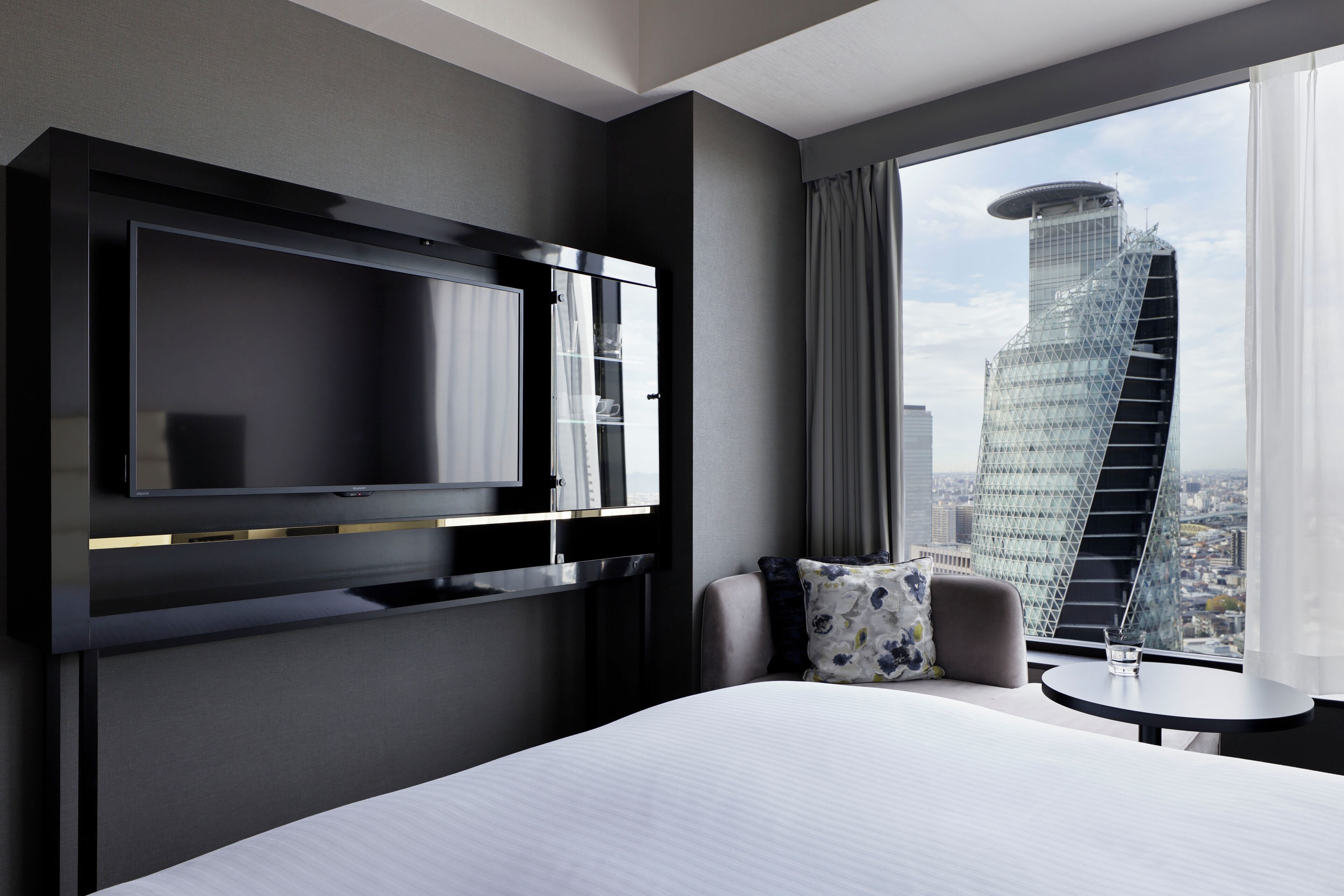Ein modernes Hotelzimmer mit einem großen Bett, einem flachbildfernseher an der Wand, einem kleinen runden Tisch und Blick auf eine Stadtlandschaft durch das Fenster.