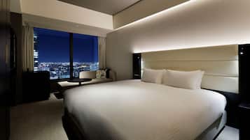 A hotel room with a large bed, a desk, a chair, and a view of a cityscape at night.