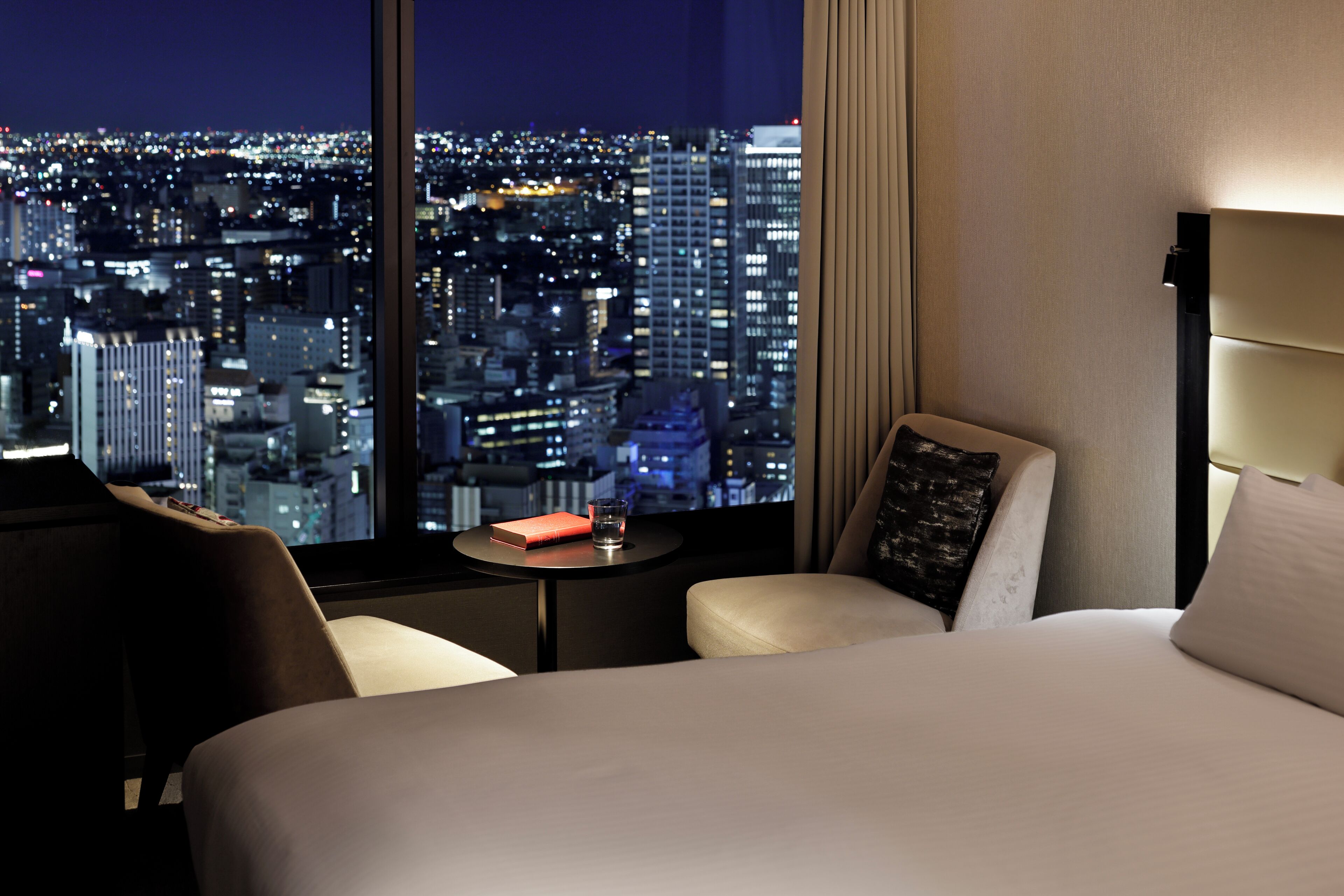 Camera d'hotel con una grande finestra che offre vista sulla città di notte, un letto, una sedia e un piccolo tavolo con un libro e un bicchiere.