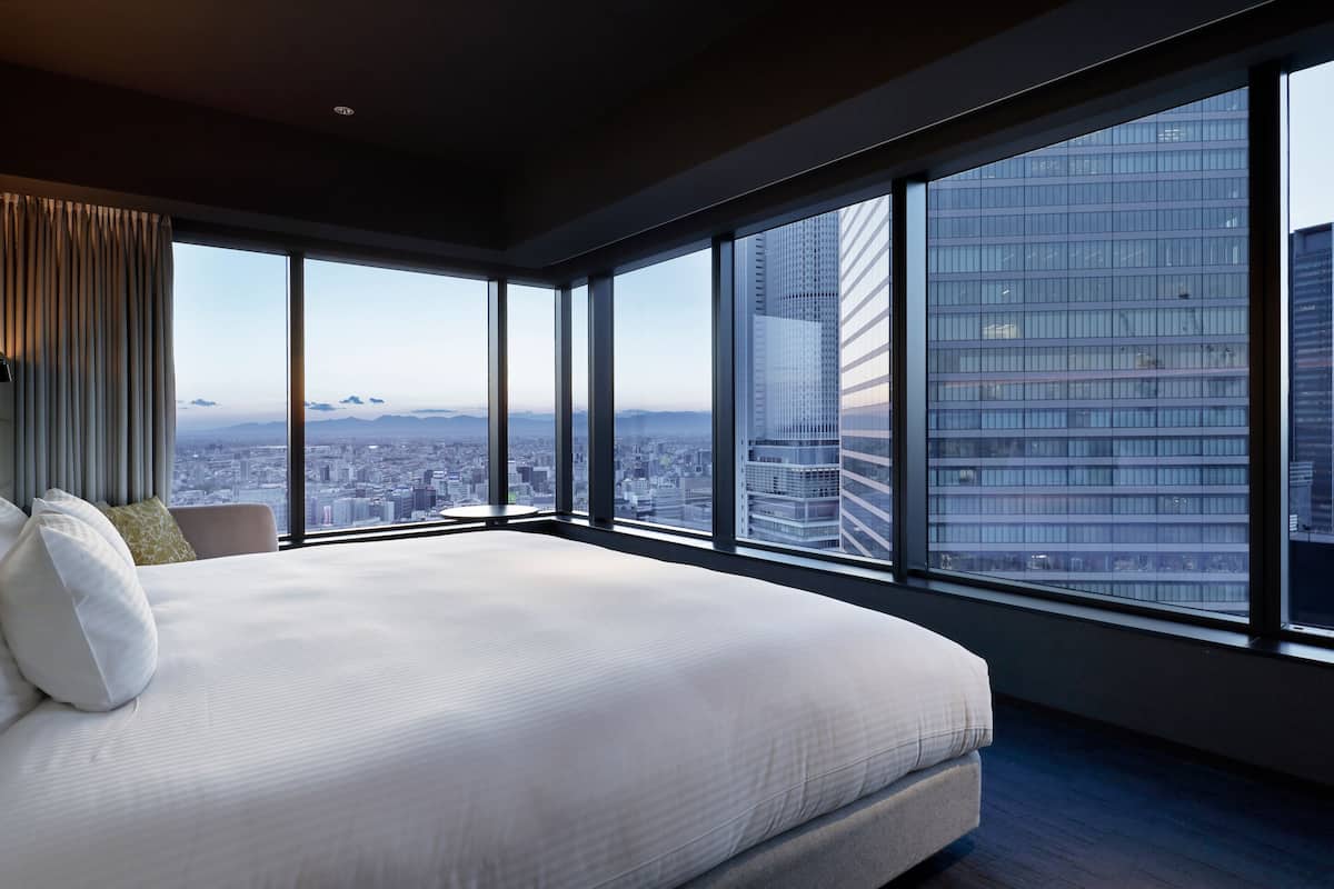 Une chambre moderne avec un grand lit, une vue sur le paysage urbain à travers de grandes fenêtres du sol au plafond, et une silhouette de la ville en arrière-plan.