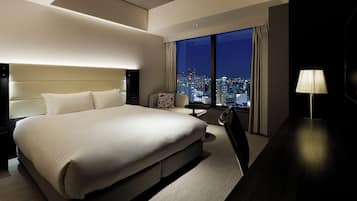 A modern hotel room with a large bed, a small table, a chair, and a view of a cityscape at night.