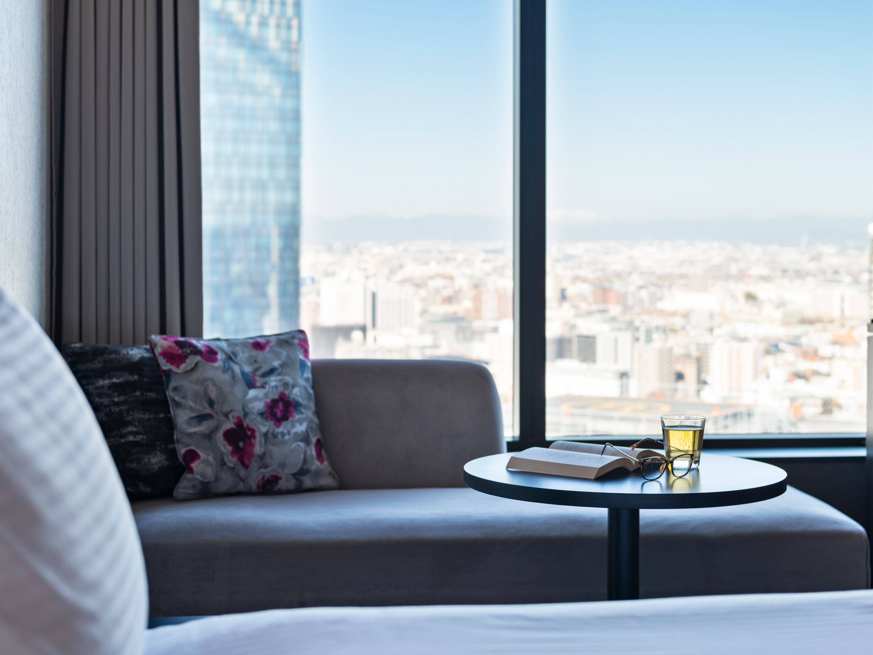 Una habitación de hotel moderna con sofá, una mesa redonda y vistas de un paisaje urbano a través de amplios ventanales.