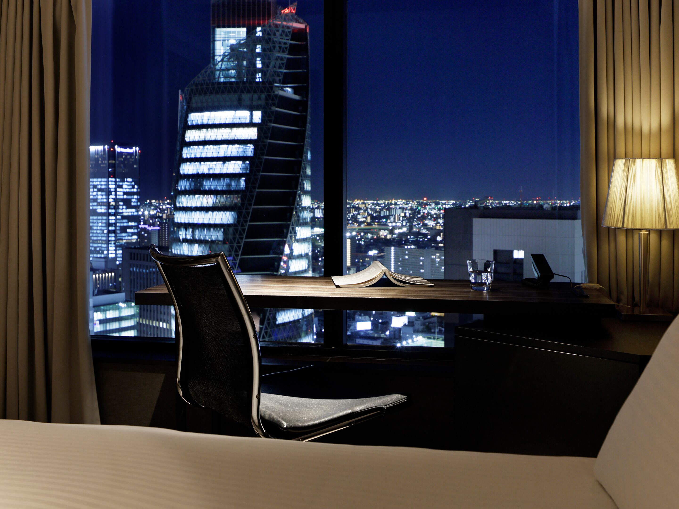 A hotel room with a view of a cityscape at night, a desk with a laptop, a chair, and an open book.