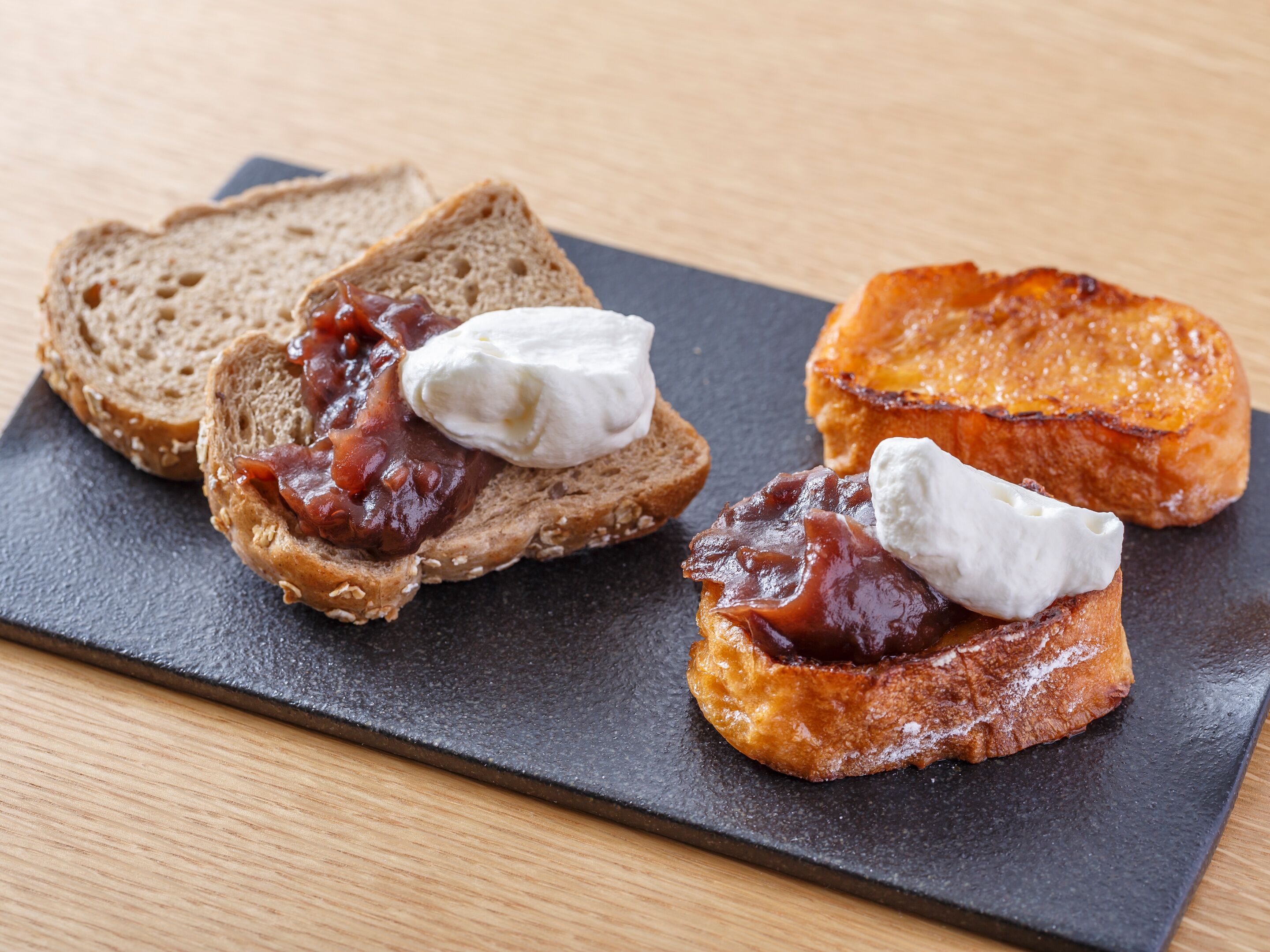 A black slate serving board with three pieces of toast topped with jam and cream cheese.