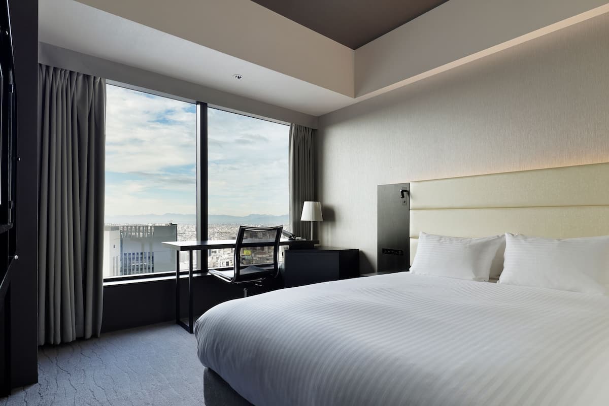 Une chambre d’hôtel avec un grand lit, un bureau, une chaise et une vue sur le paysage urbain grâce à de grandes fenêtres.