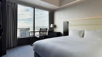 A hotel room with a large bed, a desk, a chair, and a view of the cityscape through large windows.
