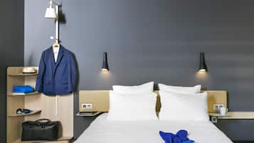 A hotel room with a neatly made bed, a hanging coat rack, a bedside table with a lamp, and a shelf with a suitcase and a blue pillow.
