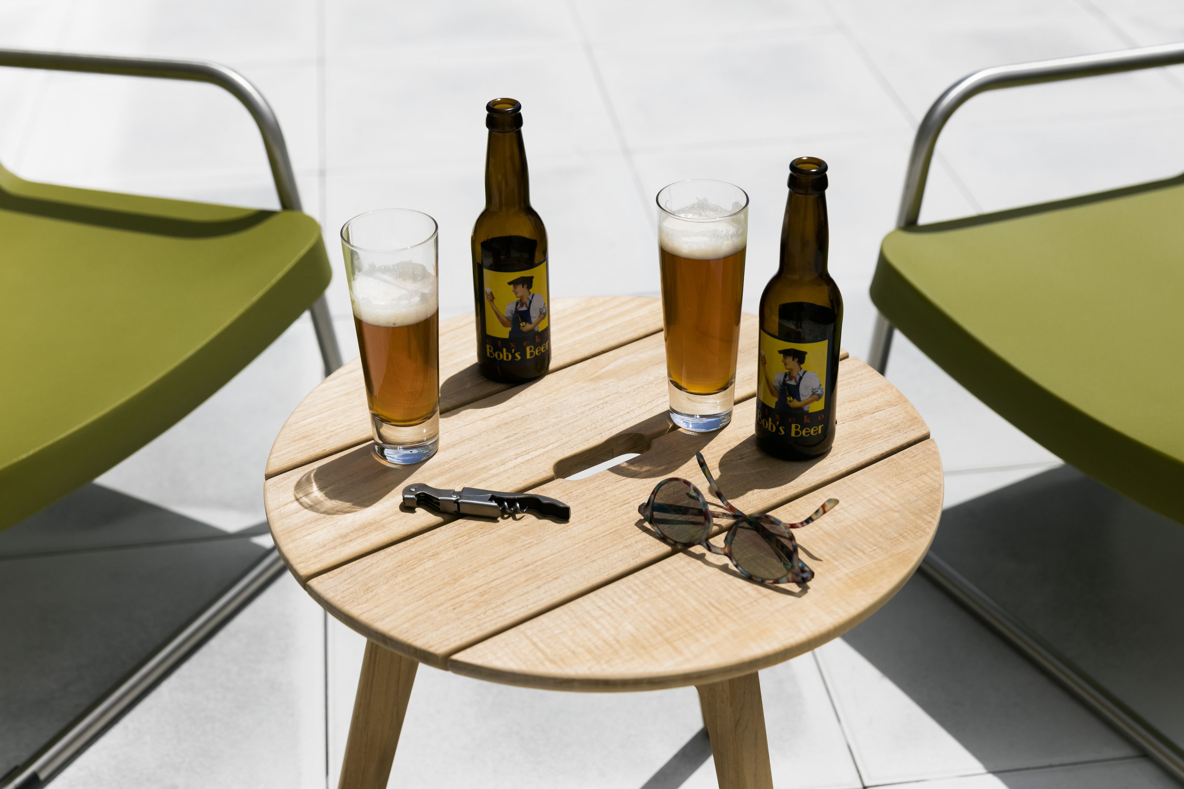 Uma mesa de madeira redonda com duas garrafas de cerveja, dois copos de cerveja, um par de óculos de sol e um abridor de garrafa.