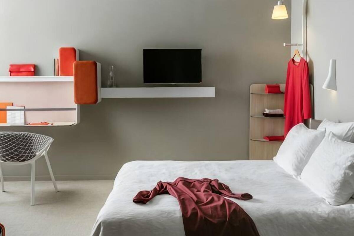 Moderni hotellihuone, jossa on sänky, työpöytä tuolilla, televisio ja punainen takki, joka on ripustettu seinälle.