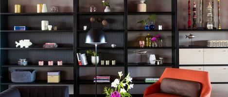 A modern living room with a black sofa, a red armchair, and a coffee table. There are shelves with decorative items and a hanging light fixture.