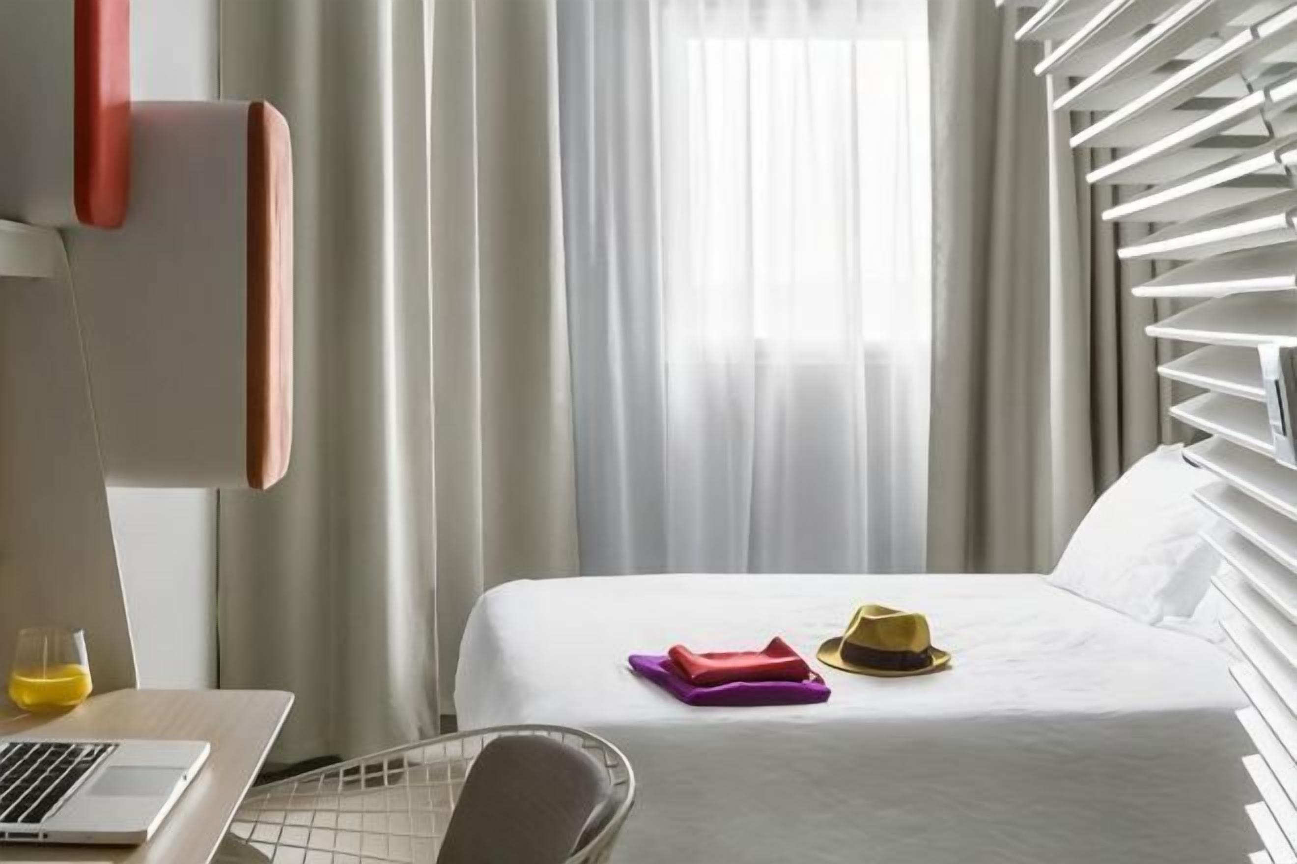 Una habitación de hotel con una cama, una computadora portátil, una silla y un ventanal con cortinas.