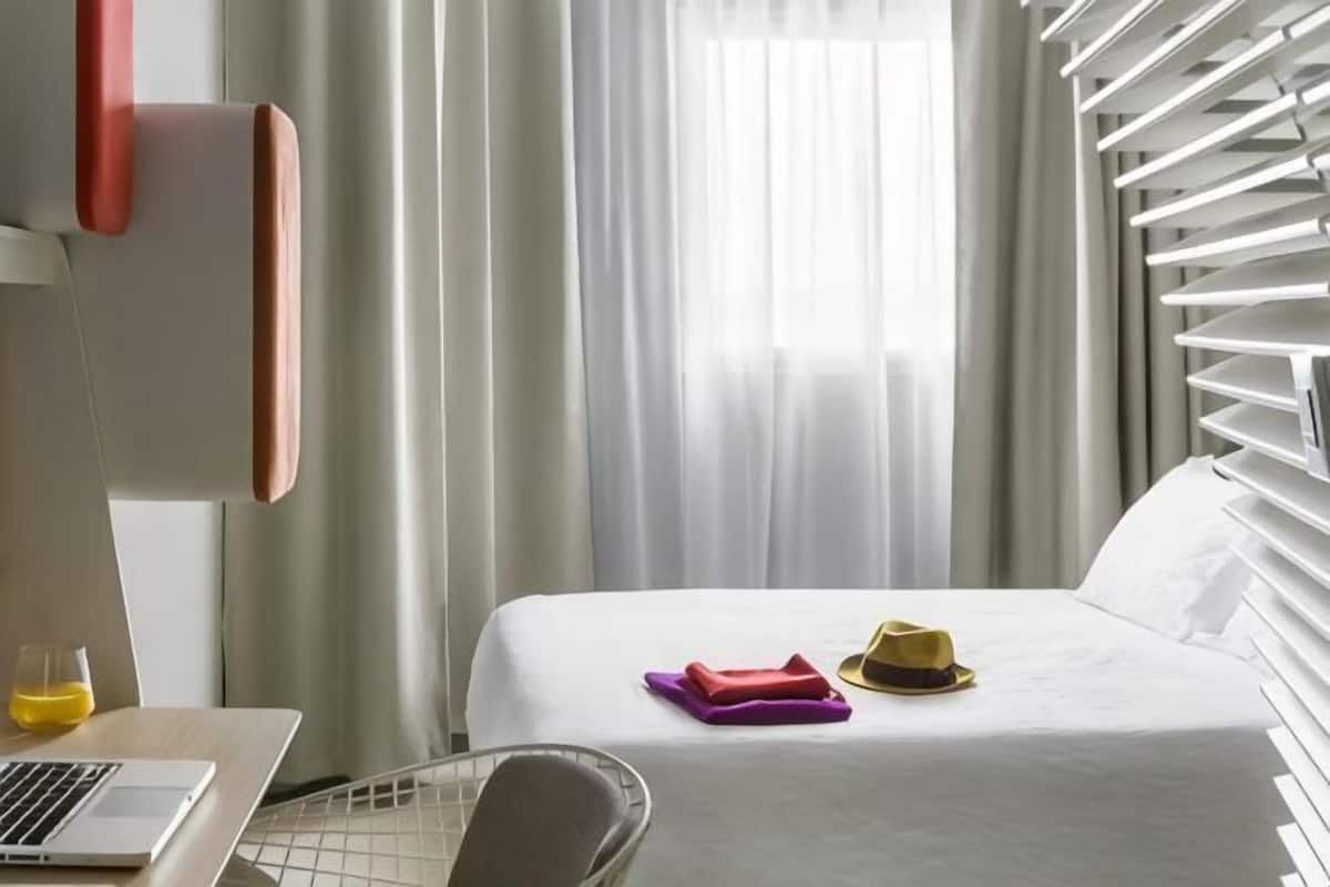 Una habitación de hotel con una cama, una computadora portátil, una silla y un ventanal con cortinas.