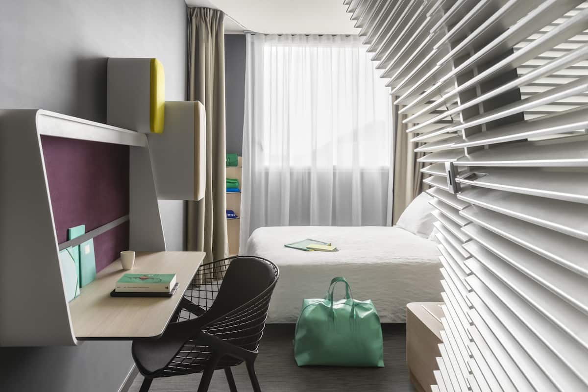 Moderni hotellihuone, jossa on sänky, työpöytä, tuoli, vihreä laukku ja ikkuna, jossa on sälekaihtimet.